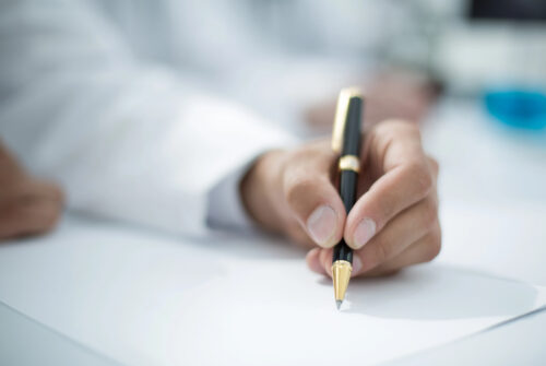Close-up of a hand in a white coat writing with a gold-and-black fountain pen on paper.