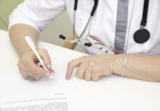 Medical professional signing a document at a white desk, with stethoscope around the neck.