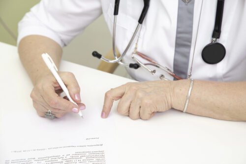 Medical professional signing a document at a white desk, with stethoscope around the neck.