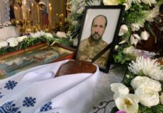 Framed portrait of a bearded man in a military uniform surrounded by white flowers and lit candles at a funeral display.