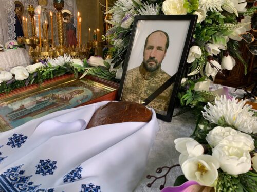 Framed portrait of a bearded man in a military uniform surrounded by white flowers and lit candles at a funeral display.