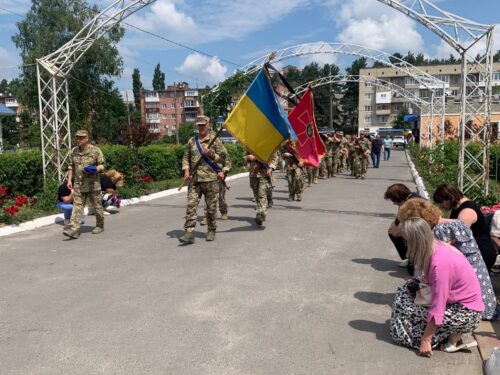Soldiers marching in a parade under a metal arch, carrying Ukrainian and red flags; spectators bow at the roadside.
