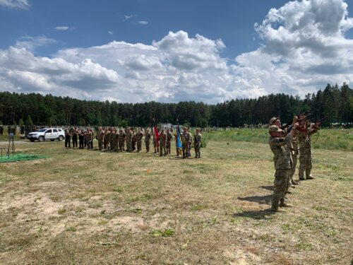 Line of soldiers in camouflage salute on a sunny grassy field with a central flag and trees in the background.