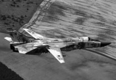 Camouflaged military fighter jet flying low over a dusty landscape, seen from above in flight.