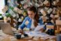 Woman working at a cluttered desk with a laptop and papers while money flies around her on all sides.