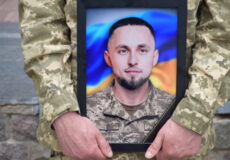 Framed portrait of a soldier in camouflage being held by another uniformed person, with a Ukrainian flag backdrop.