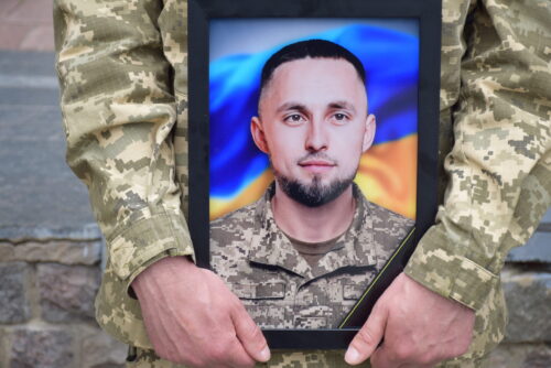 Framed portrait of a soldier in camouflage being held by another uniformed person, with a Ukrainian flag backdrop.