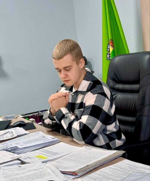 Man in a plaid shirt sits at a desk with scattered documents, hands clasped and eyes closed.