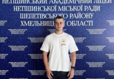 Young man in a white T-shirt stands in front of a blue backdrop covered with Ukrainian text the far wall.