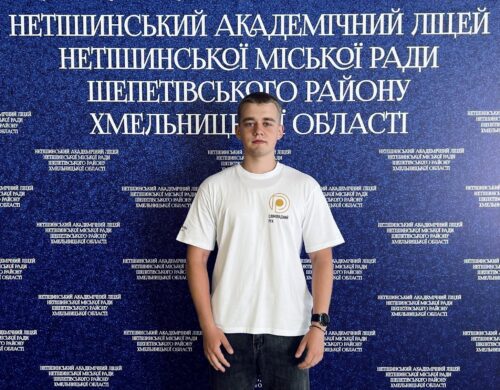 Young man in a white T-shirt stands in front of a blue backdrop covered with Ukrainian text the far wall.