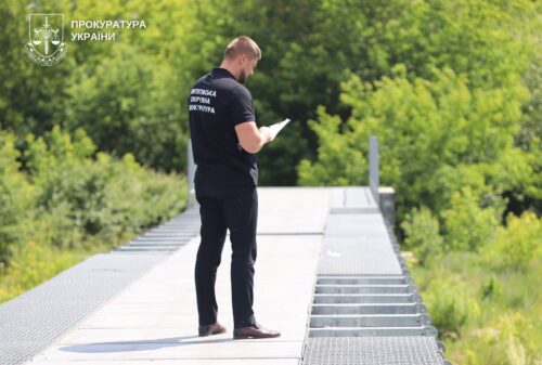 Uniformed Ukrainian prosecutor from the Prosecutor's Office standing on a metal pedestrian bridge, taking notes on a clipboard.
