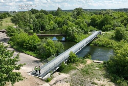 Pedestrian bridge over a river in a green park; three workers in high-visibility vests near the near end, logo in the top-left corner.