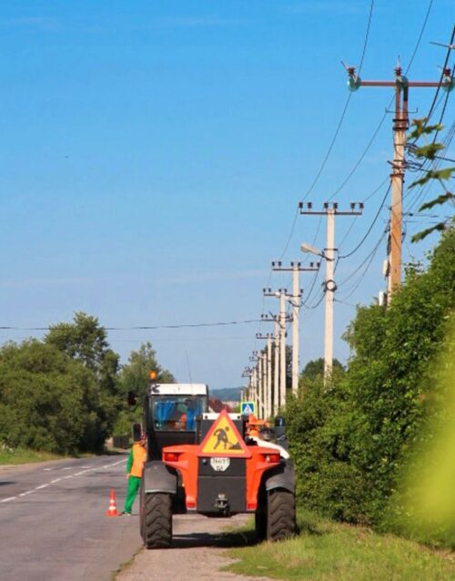 Orange road roller with a caution sign and a worker in high-visibility clothing on a rural road, cones nearby and power poles lining the street.