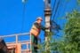 Utility worker in orange safety vest and helmet on an elevated lift repairing power lines on a utility pole against a blue sky.