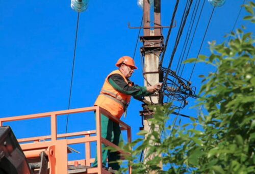 Utility worker in orange safety vest and helmet on an elevated lift repairing power lines on a utility pole against a blue sky.