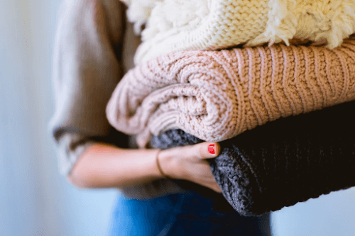 Person holding a stack of folded knitted sweaters in cream, pink, and gray colors.