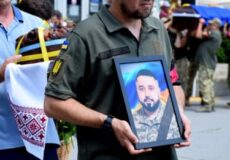 Man in a military uniform carries a framed portrait of a bearded man in camouflage during a memorial or funeral procession, with a wreath and onlookers in the background.