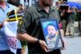 Man in a military uniform carries a framed portrait of a bearded man in camouflage during a memorial or funeral procession, with a wreath and onlookers in the background.
