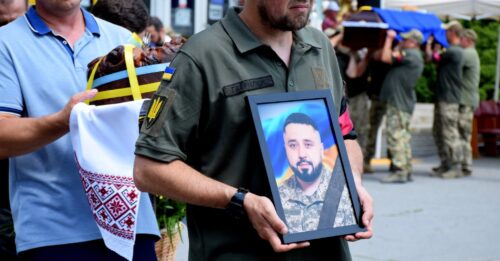 Man in a military uniform carries a framed portrait of a bearded man in camouflage during a memorial or funeral procession, with a wreath and onlookers in the background.