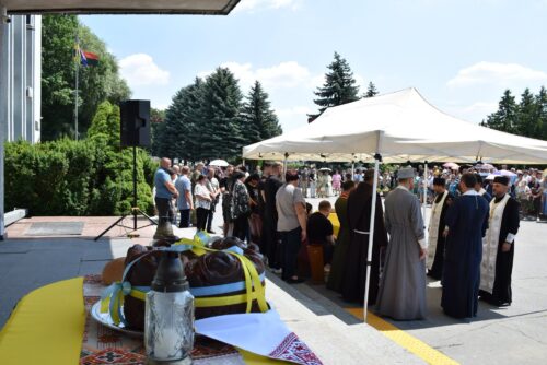 Outdoor ceremony with a large decorated cake on a table in the foreground, a crowd under a white canopy, and clergy in robes in the background.