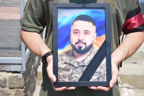 Person in camouflage uniform holding a framed portrait of a bearded man in military attire.