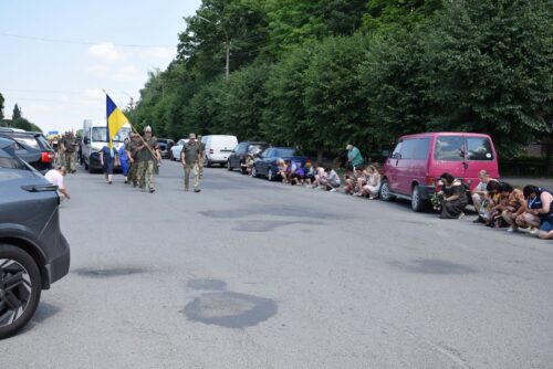 Soldiers march down a residential street in a parade; spectators sit along the curb on both sides, and a yellow-and-blue flag leads the procession.