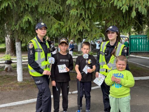 Two female police officers in high-visibility vests stand with three children outdoors, distributing pamphlets at a community outreach event.