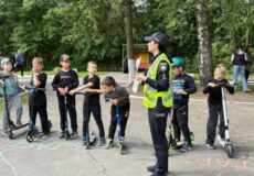 Police officer in a reflective vest giving scooter safety instruction to a line of kids in a park with chalk drawings on the pavement.
