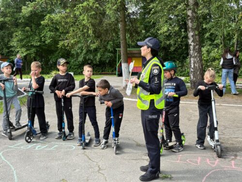 Police officer in a reflective vest giving scooter safety instruction to a line of kids in a park with chalk drawings on the pavement.