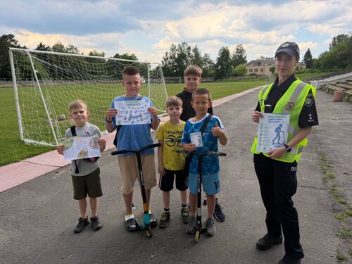 Group of six young boys posing with certificates on a paved track near a soccer goal, with one boy on a scooter and another in front holding up his certificate, a safety officer in a high-visibility vest stands to the right holding a certificate.