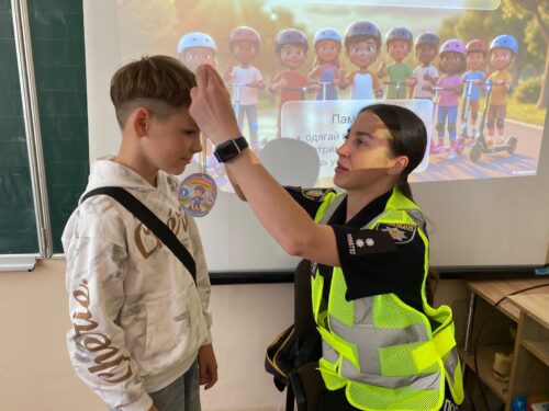 Female officer in a reflective vest takes a boy’s forehead temperature with a handheld thermometer in a classroom.