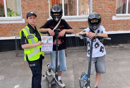 Police officer in a high-visibility vest standing with two kids on scooters wearing helmets, holding a safety flyer outside a brick building.