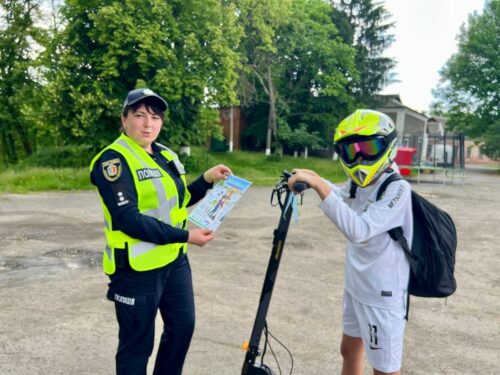 Police officer in a neon vest hands a safety flyer to a rider on an electric scooter wearing a helmet outdoors.