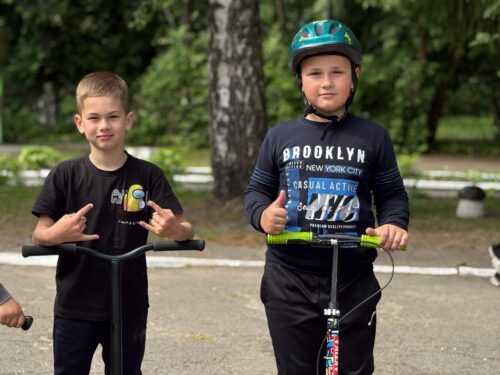 Two boys with kick scooters in a park; the boy on the left shows a peace sign, the boy on the right wears a helmet and gives a thumbs up.