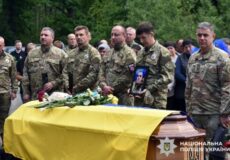 Ukrainian soldiers stand around a coffin draped with a yellow flag, with flowers on top, as civilians look on during a funeral; a soldier holds a portrait of the deceased.