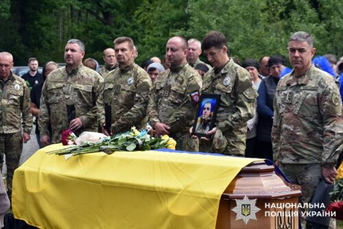 Ukrainian soldiers stand around a coffin draped with a yellow flag, with flowers on top, as civilians look on during a funeral; a soldier holds a portrait of the deceased.