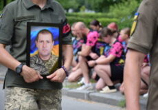 Soldier in camouflage holds framed portrait of a uniformed man at a memorial gathering.