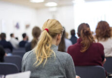 Rear view of a conference audience with a blonde woman in a grey blazer seated in the foreground.