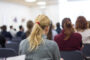 Rear view of a conference audience with a blonde woman in a grey blazer seated in the foreground.
