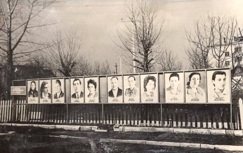 Row of portrait posters mounted on boards along a fence in a winter scene, likely a memorial display with bare trees in the background.