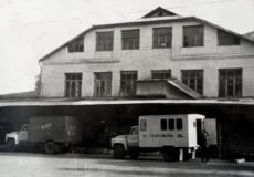 Black-and-white photograph of a brick two-story warehouse with loading docks and several trucks parked in front, with a few people near a small white service truck.