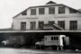 Black-and-white photograph of a brick two-story warehouse with loading docks and several trucks parked in front, with a few people near a small white service truck.