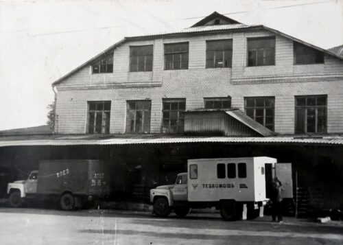 Black-and-white photograph of a brick two-story warehouse with loading docks and several trucks parked in front, with a few people near a small white service truck.