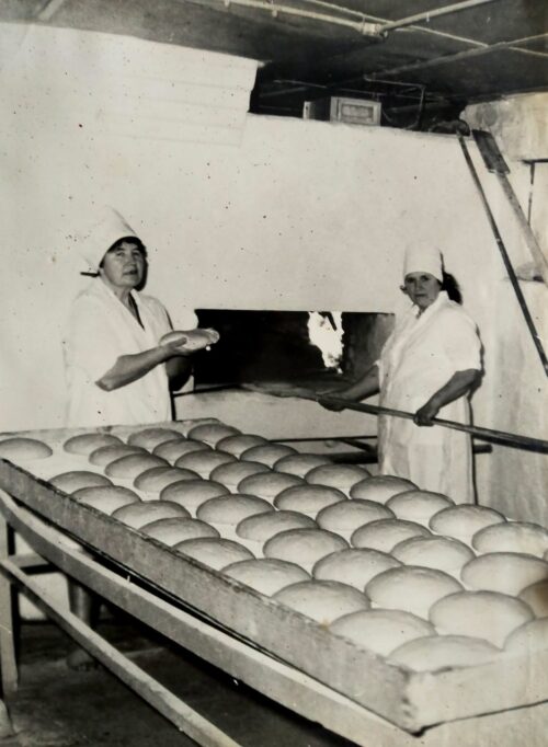 Two women in white uniforms and caps stand beside a long tray filled with neatly arranged eggs, inspecting or loading them into a large bakery oven.