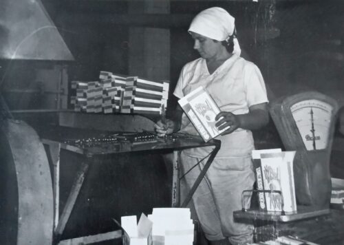 Black-and-white photo of a female factory worker in a hairnet and apron, arranging stacked chocolate bars on a table near a large scale, holding a box of chocolates.