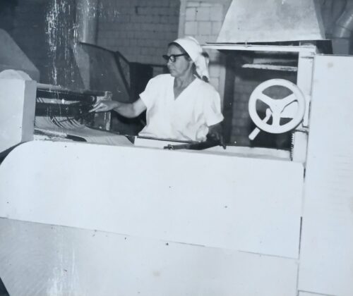 Worker in white uniform and headscarf operating a large industrial machine with a side wheel and control panel.