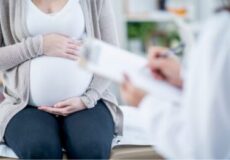 Pregnant woman sitting on an exam table while a clinician shows her a document or form nearby