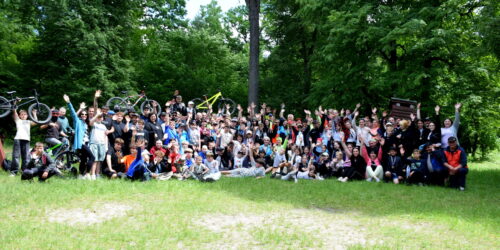 Large group of people in a park posing for a photo; several bicycles are held aloft above their heads in celebration.