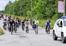 Large group of cyclists riding together on a sunny road, with a police SUV leading on the right.