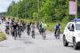 Large group of cyclists riding together on a sunny road, with a police SUV leading on the right.